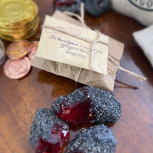 May include: A small brown paper package tied with twine and labeled "The Head Goblin & Gringotts Bank".  The package is sitting on a wooden surface with a pile of gold coins and three red and black gemstone-like objects.