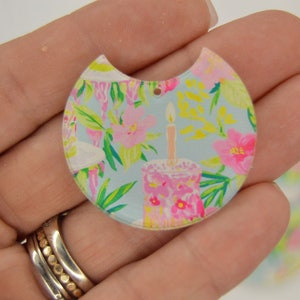May include: A light blue, floral patterned, half-circle acrylic charm with a hole for hanging. The charm features a pink and white cake with a lit candle.