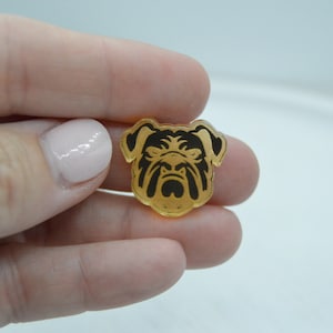 Pu&ograve; includere: Un ciondolo in acrilico color oro con una silhouette nera di una testa di bulldog.