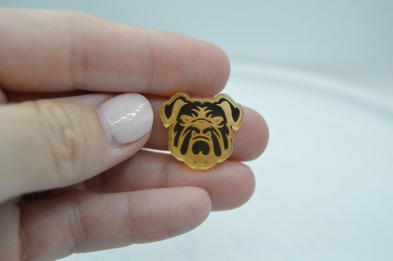 Pu&ograve; includere: Un ciondolo in acrilico color oro con una silhouette nera di una testa di bulldog.