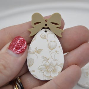 Könnte beinhalten: Ein weißer eiförmiger Anhänger mit einem Blumenmuster und einer braunen Schleife oben. Der Anhänger ist aus Holz und hat einen rustikalen, Vintage-Look.