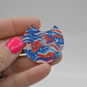 May include: A red, white, and blue patterned resin cabochon with a crescent moon shape. The pattern features wavy lines and stars.