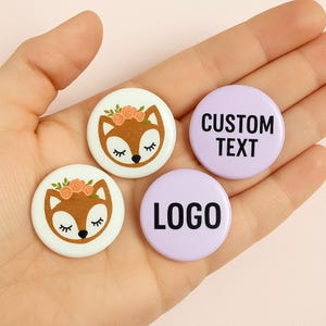 May include: Three round button pins. One features a cartoon fox with a flower crown, another says "CUSTOM TEXT", and the last one says "LOGO".