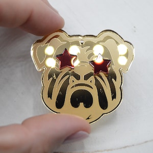 Pu&ograve; includere: Spilla a forma di bulldog dorata con occhi a forma di stella rossa. La spilla ha una finitura lucida e metallica.