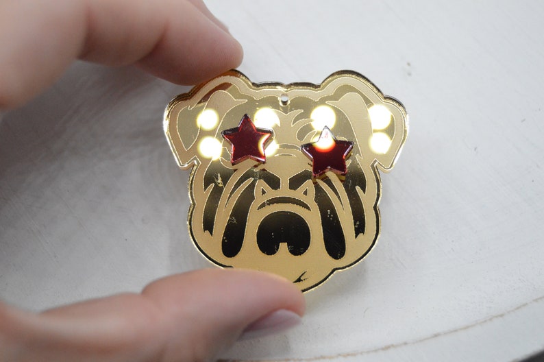 Pu&ograve; includere: Spilla a forma di bulldog dorata con occhi a forma di stella rossa. La spilla ha una finitura lucida e metallica.