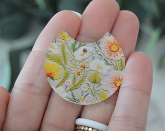 Può includere: Un ciondolo bianco, rotondo, a metà cerchio con un motivo floreale. Il ciondolo ha un foro nella parte superiore per essere attaccato a una collana o un braccialetto. I fiori sono ricamati in filo giallo, arancione e bianco.
