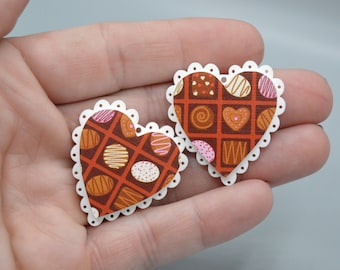 Può includere: Due ritagli di carta a forma di cuore con bordo smerlato. I cuori presentano un design a scatola di cioccolatini con vari cioccolatini marroni, bianchi, rosa e arancioni. I cuori sono tenuti in una mano.