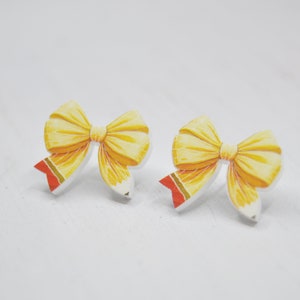 May include: A pair of yellow bow earrings with red and gold accents. The bows are made of a lightweight material and have a glossy finish.