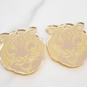 May include: Two gold-colored acrylic ornaments shaped like tiger faces. The tiger faces are outlined in gold and have a light brown background.