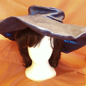 Op de afbeelding: Een grote, breedgerande hoed gemaakt van donkerblauwe stof met een glanzende, bijna metallic afwerking. De hoed wordt gedragen op een mannequin met bruin haar.