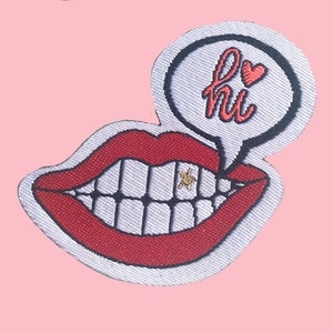 Può includere: Due toppe ricamate raffiguranti una bocca sorridente con labbra rosse e denti bianchi. Una nuvoletta sopra la bocca dice "hi" con un cuore. Un dente ha una stella d'oro. Su sfondo rosa.