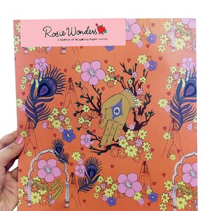 Peut inclure: Papier d'emballage orange avec un motif r&eacute;p&eacute;titif de fleurs roses, de plumes de paon, de mains en position de pri&egrave;re et de petits c&oelig;urs. Le texte "Rosie Wonders" est imprim&eacute; en rose en haut du papier.