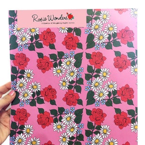 May include: Pink wrapping paper with a repeating pattern of red roses and white daisies. The paper has the text "Rosie Wonders 3 sheets of Wrapping Paper 50cm x 70cm" printed on it.