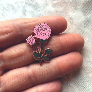 May include: A gold-tone enamel pin featuring a pink rose with green leaves. The rose has a single bud and a larger bloom.