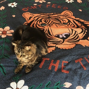 K&ouml;nnte beinhalten: Eine braune getigerte Katze sitzt auf einer blauen und gr&uuml;nen Wurfdecke mit einem Tigergesicht und dem Text "Don't Feed The Tiger". Die Decke hat ein Blumenmuster mit wei&szlig;en und rosa Bl&uuml;ten.