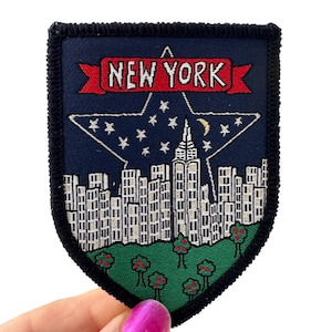 May include: Embroidered patch featuring a shield design with a New York City theme. The patch has a navy blue background with a white star and cityscape design. The text "NEW YORK" is displayed in red.