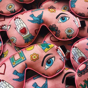 May include: A close-up of a pile of pink sleep masks with various colorful designs, including a cactus, a bird, an eye, a hand, and a heart. The masks are made of soft fabric and have a black elastic band.