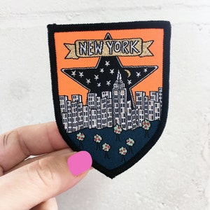 May include: An orange and black embroidered patch featuring a star with the text "NEW YORK". The star contains a cityscape and a crescent moon at the top. The cityscape is surrounded by small trees with red flowers.