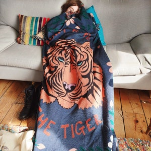 K&ouml;nnte beinhalten: Eine blaue und orangefarbene Wolldecke mit einem Tigergesicht und dem Aufdruck "Don't Be The Tiger". Die Decke ist aus weichem, flauschigem Material und hat ein Blumenmuster.