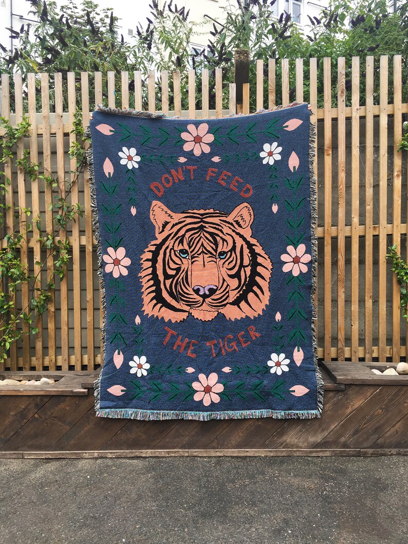 K&ouml;nnte beinhalten: Eine blaue gewebte Wolldecke mit einem Tigergesicht und dem Text "Don't Feed The Tiger" in Orange und Wei&szlig;. Die Decke hat einen floralen Rand mit rosa und wei&szlig;en Blumen.