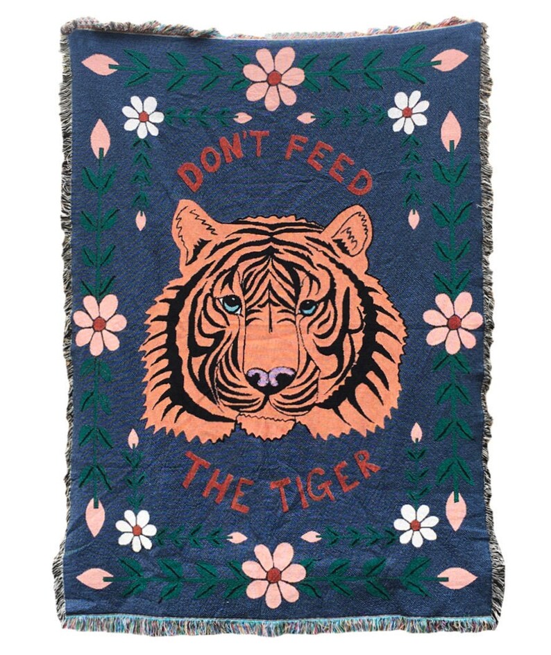 K&ouml;nnte beinhalten: Eine blaue gewebte Wolldecke mit einem Tigergesicht in der Mitte. Der Tiger ist orange und schwarz mit blauen Augen. Der Text "Don't Feed The Tiger" ist in roten und wei&szlig;en Buchstaben &uuml;ber dem Tiger geschrieben. Die Decke ist von einem Rand aus gr&uuml;nen Bl&auml;ttern und rosa und wei&szlig;en Blumen umgeben.