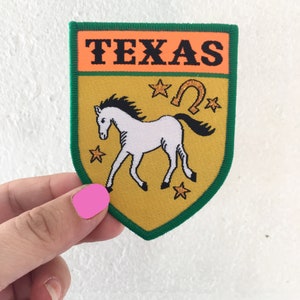 Peut inclure: Un écusson brodé jaune et vert avec le mot "TEXAS" en lettres noires sur fond orange. L'écusson représente un cheval blanc avec un fer à cheval et des étoiles en paillettes dorées.