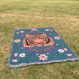 K&ouml;nnte beinhalten: Eine blaue gewebte Wolldecke mit einem Tigergesicht und dem Text "Don't Feed The Tiger" in Rot. Die Decke hat einen floralen Rand mit rosa und wei&szlig;en Blumen.