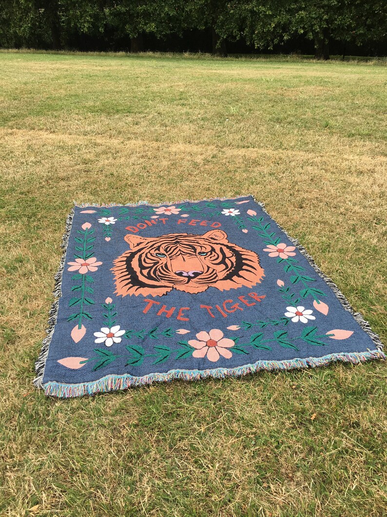 K&ouml;nnte beinhalten: Eine blaue gewebte Wolldecke mit einem Tigergesicht und dem Text "Don't Feed The Tiger" in Rot. Die Decke hat einen floralen Rand mit rosa und wei&szlig;en Blumen.