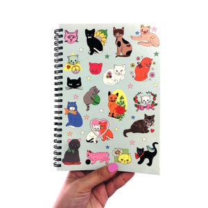 May include: A spiral bound notebook with a light blue cover featuring a variety of colorful cat illustrations. The cats are depicted in various poses and expressions, including sleeping, playing, and looking playful. The notebook is perfect for jotting down notes, ideas, or sketches.