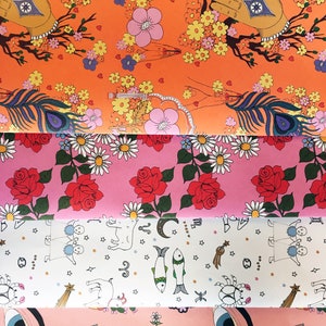 May include: Four different patterned wrapping papers. The top paper is orange with pink and yellow flowers, a peacock feather, and a hand with a star. The second paper is pink with red roses and white daisies. The third paper is white with black stars and zodiac symbols. The bottom paper is pink with a green cactus, a yellow sun, a white daisy, and a blue eye.