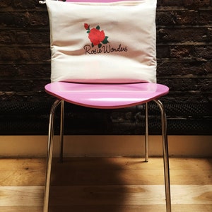 K&ouml;nnte beinhalten: Ein wei&szlig;es Kissen aus Canvas mit einem roten Rosen- und gr&uuml;nen Bl&auml;tterdesign und dem Text "Rosie Wonders" auf einem rosa Stuhl.