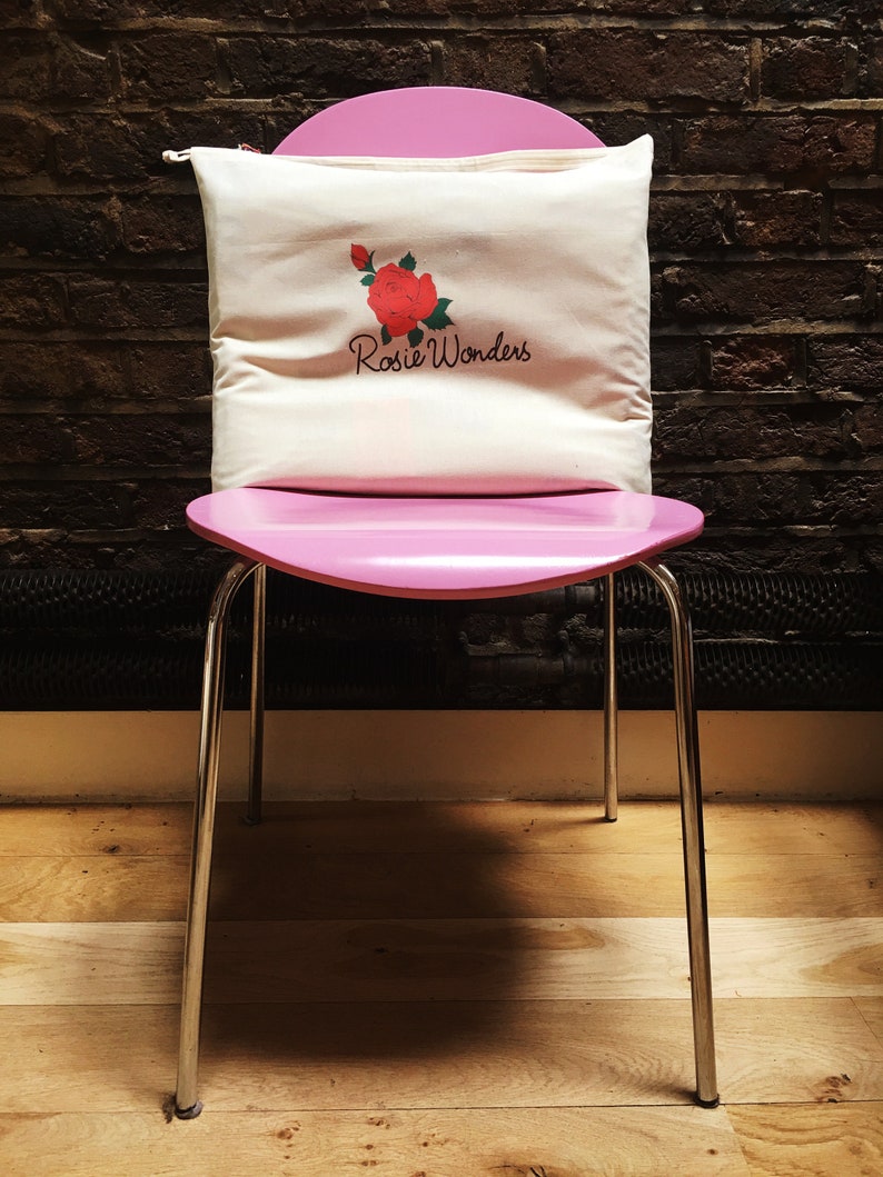 K&ouml;nnte beinhalten: Ein wei&szlig;es Kissen aus Canvas mit einem roten Rosen- und gr&uuml;nen Bl&auml;tterdesign und dem Text "Rosie Wonders" auf einem rosa Stuhl.