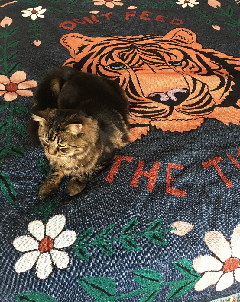 K&ouml;nnte beinhalten: Eine braune getigerte Katze sitzt auf einer blauen und gr&uuml;nen Wurfdecke mit einem Tigergesicht und dem Text "Don't Feed The Tiger". Die Decke hat ein Blumenmuster mit wei&szlig;en und rosa Bl&uuml;ten.