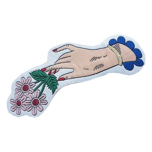 May include: An embroidered iron-on patch featuring a hand with red nail polish holding a bouquet of pink flowers with green leaves. The hand is wearing a gold bracelet and a blue cuff.