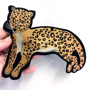 May include: An embroidered patch of a leopard, featuring brown and black fur, against a white background.