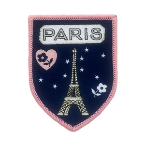May include: A navy blue fabric patch with pink stitching. The patch features a gold embroidered Eiffel Tower, a pink heart with a white flower, and a white flower with a green stem. The text "PARIS" is embroidered in white with a gold outline.