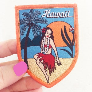 Op de afbeelding: Een geborduurde patch in vintage stijl met een hula-danseres in een rode rok en een bloem in haar haar, een palmboom en een zonsondergang. De patch is omgeven door een oranje rand en het woord "Hawaii" is geborduurd in wit op een blauwe achtergrond.