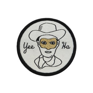 May include: A white embroidered patch with a black outline of a person wearing a cowboy hat and a gold mask. The text "Yee Ha" is embroidered on the patch.