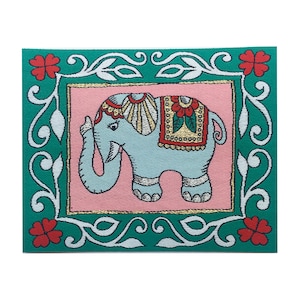 May include: A teal blue square with a white and gold frame. Inside the frame is a pink background with a light blue elephant with a red, gold, and white patterned saddle. The elephant is decorated with gold trim and red flowers. The frame is decorated with white floral designs and red flowers.