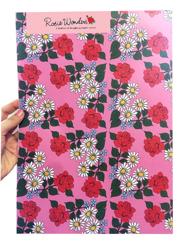 May include: Pink wrapping paper with a repeating pattern of red roses and white daisies. The paper has the text "Rosie Wonders 3 sheets of Wrapping Paper 50cm x 70cm" printed on it.
