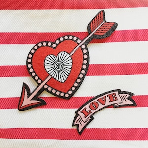 Puede incluir: Un corazón rojo con un corazón blanco en su interior está atravesado por una flecha negra y roja. Debajo del corazón hay una pancarta rosa con la palabra "LOVE" en letras rojas.