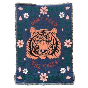 K&ouml;nnte beinhalten: Eine gewebte Wolldecke mit blauem Hintergrund und einem pinkfarbenen und orangen Tigergesicht in der Mitte. Der Text "Don't Feed The Tiger" ist in roten und wei&szlig;en Buchstaben &uuml;ber dem Tiger geschrieben. Die Decke ist mit gr&uuml;nen Ranken und pinkfarbenen und wei&szlig;en Blumen verziert.