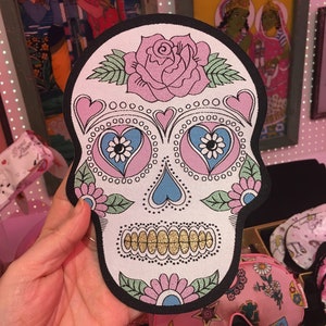 Può includere: Un teschio di zucchero rosa e blu con una rosa e disegni floreali. Il teschio è fatto di tessuto e ha un bordo nero.