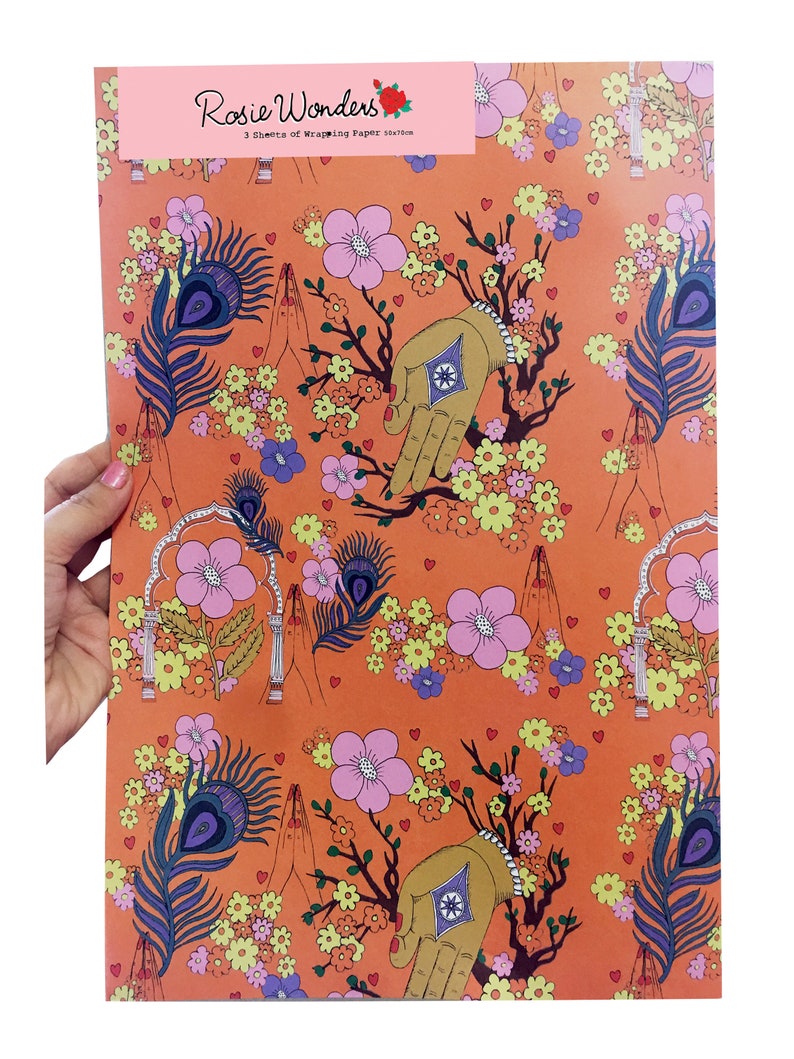 Peut inclure: Papier d'emballage orange avec un motif r&eacute;p&eacute;titif de fleurs roses, de plumes de paon, de mains en position de pri&egrave;re et de petits c&oelig;urs. Le texte "Rosie Wonders" est imprim&eacute; en rose en haut du papier.