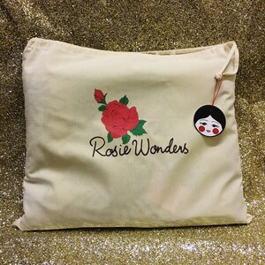 K&ouml;nnte beinhalten: Eine wei&szlig;e Canvas-Tragetasche mit einem Design aus einer roten Rose und gr&uuml;nen Bl&auml;ttern. Die Tasche ist mit dem Text "Rosie Wonders" bedruckt. Ein kleines rotes und schwarzes Puppen-Gesichts-Etikett ist an der Tasche befestigt.