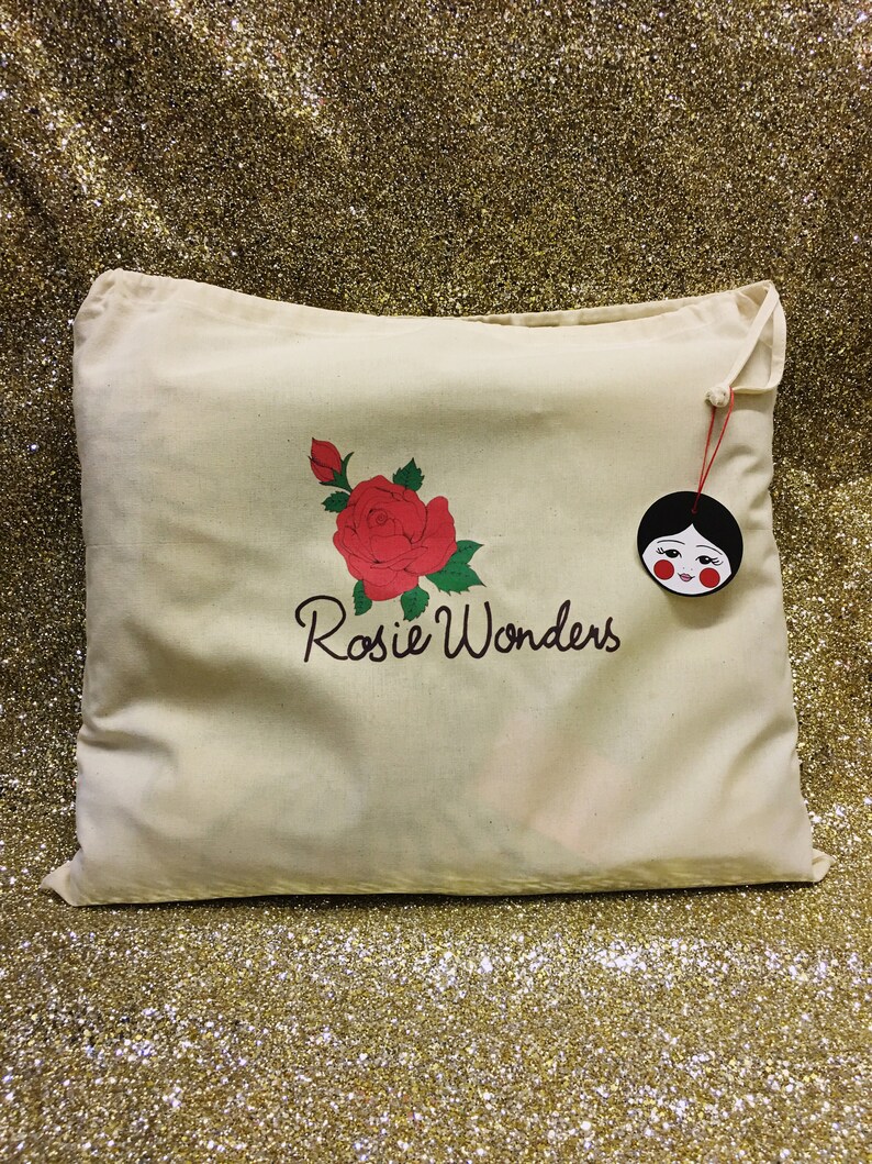 K&ouml;nnte beinhalten: Eine wei&szlig;e Canvas-Tragetasche mit einem Design aus einer roten Rose und gr&uuml;nen Bl&auml;ttern. Die Tasche ist mit dem Text "Rosie Wonders" bedruckt. Ein kleines rotes und schwarzes Puppen-Gesichts-Etikett ist an der Tasche befestigt.