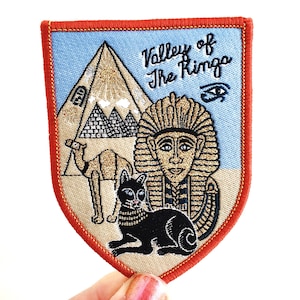 May include: A brown and orange embroidered patch with a blue background. The patch features a pyramid, a camel, a cat, and a pharaoh. The text "Valley of the Kings" is embroidered in black thread.