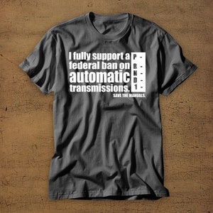 May include: A dark gray t-shirt with a white graphic that reads "I fully support a federal ban on automatic transmissions. SAVE THE MANUALS." The graphic includes a stylized gear shift pattern with the letters "P-R-N-D-1".