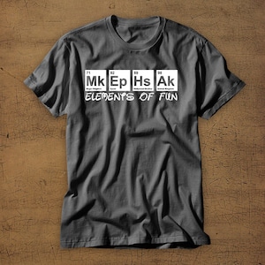 May include: A dark gray t-shirt with a white graphic of the periodic table elements. The elements are labeled with the abbreviations "Mk", "Ep", "Hs", and "Ak" and the text "Elements of Fun".