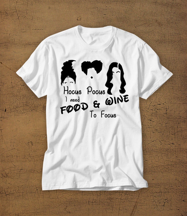 May include: White t-shirt with a black graphic of the Sanderson sisters from the movie Hocus Pocus. The text reads "Hocus Pocus I need FOOD & WINE To Focus".